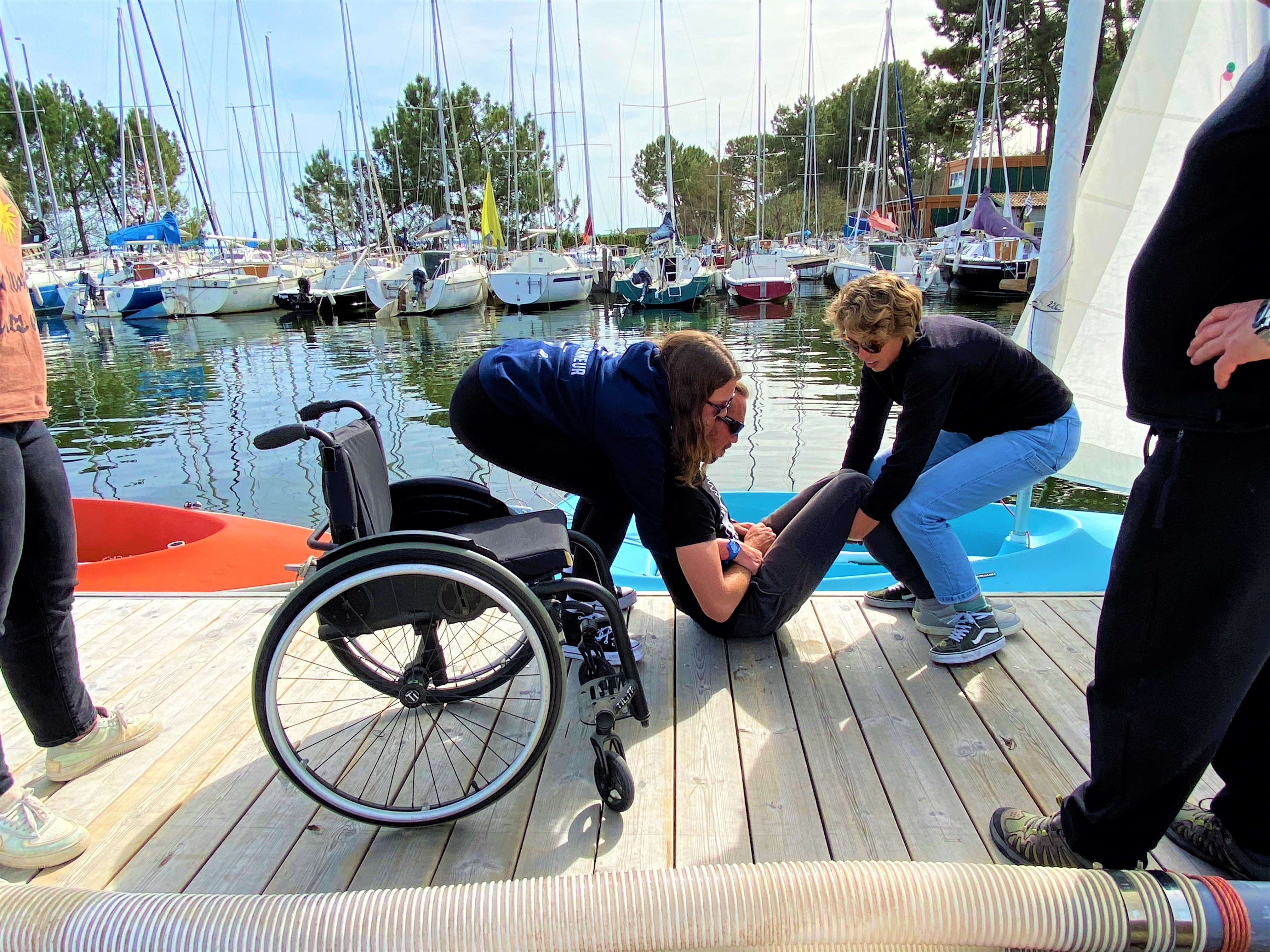 Sur un ponton, deux monitrices de voile soulèvent une personne paraplégique pour la replacer dans son fauteuil après avoir navigé